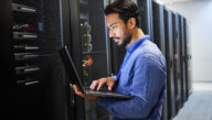 Man standing in front of a bank of computer servers, working on a laptop