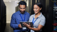 Two IT professionals working on a tablet while standing in front of a bank of servers