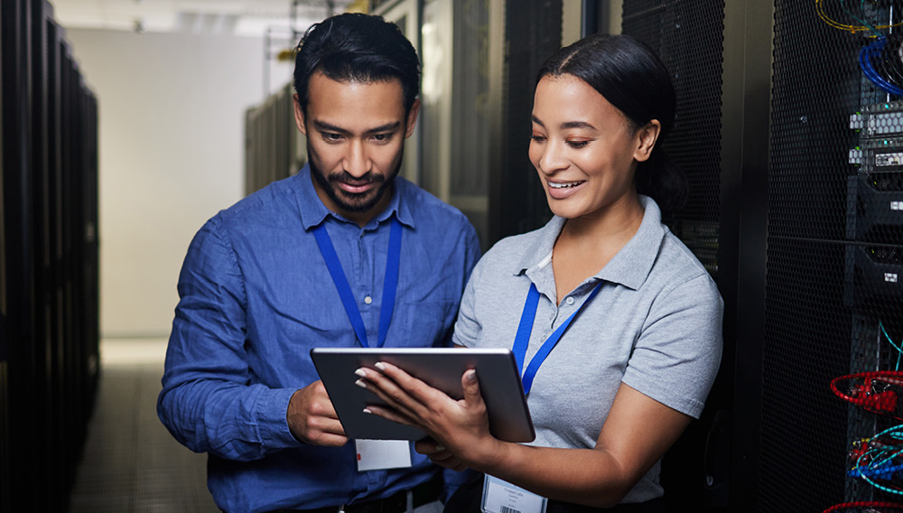 Two IT professionals working on a tablet while standing in front of a bank of servers