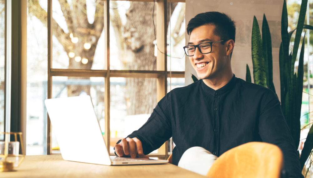 Man smiling at his laptop, seated in front of a window