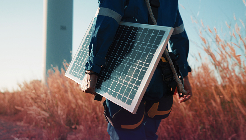 An electrical engineering technician carries a solar panel through a field.