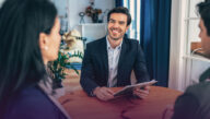 Smiling man seated behind a desk and holding a clipboard, engaging customers