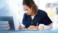 Nurse sitting at a table with a laptop, books, and a notebook, studying for the NCLEX-RN