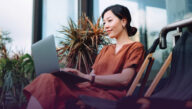 Seated, smiling woman works on her laptop in an atrium full of plants